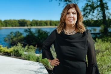 Laura Clancy stands outside on a nature trail in front of a small lake. The sky is bright blue and has no clouds.