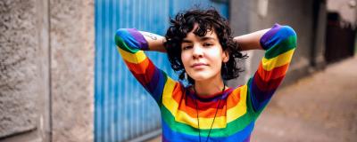 A person wearing a rainbow striped sweater stands outside, hands behind head